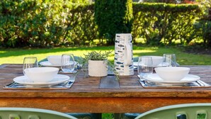 A wooden picnic table set outdoors with white plates, bowls, and glasses, surrounded by greenery.