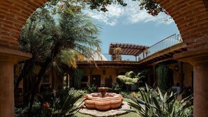 Une cour intérieure avec une fontaine centrale, entourée d’une végétation tropicale luxuriante et d’une arche en briques.