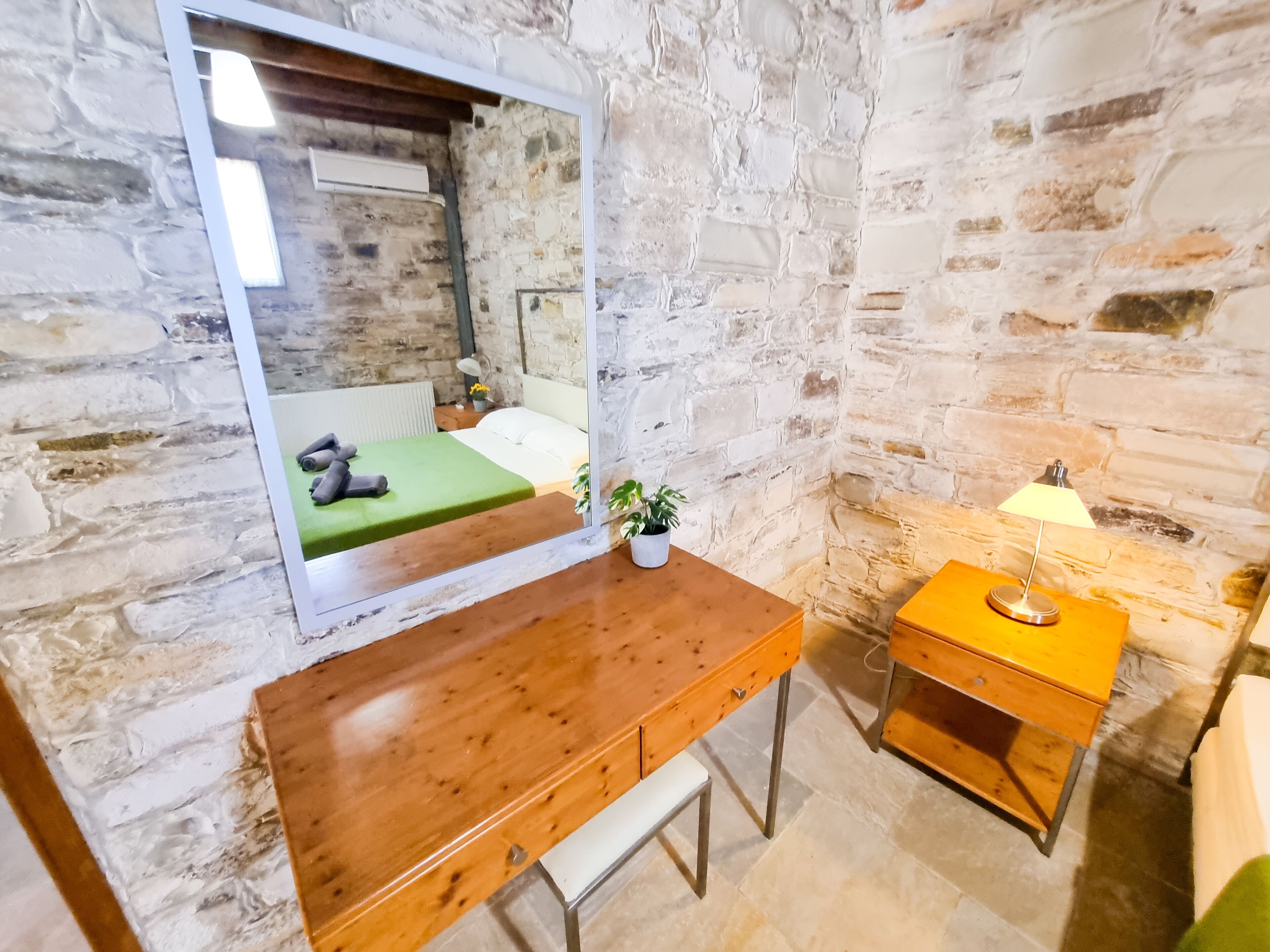 A room with a stone wall, a wooden desk, a mirror, a bedside table, and a lamp.