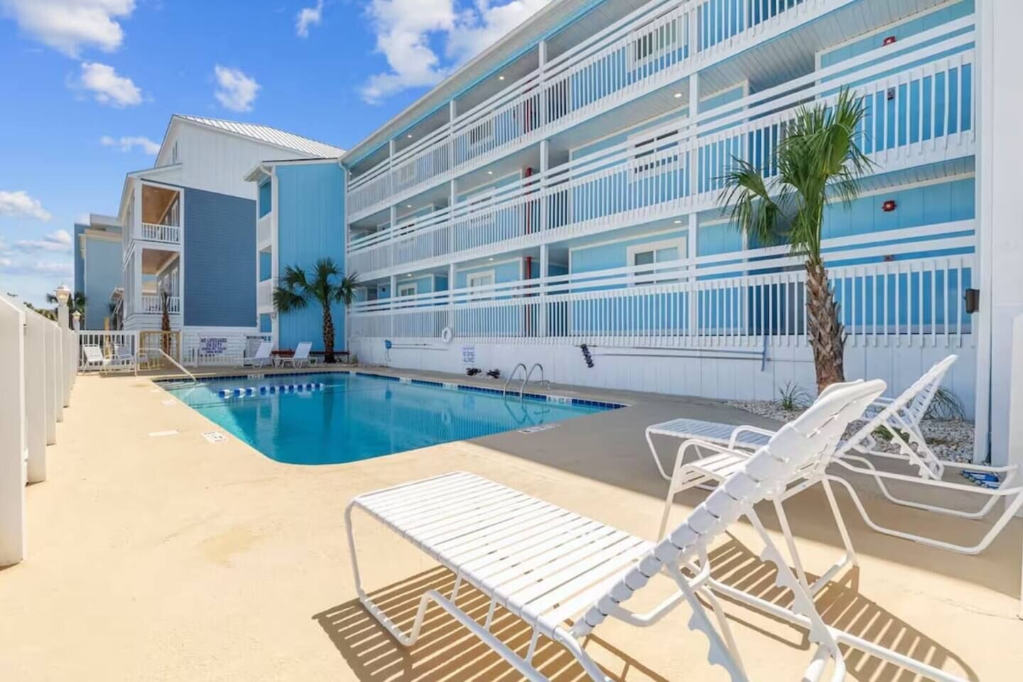 A hotel pool area with a clear blue pool, white lounge chairs, and a multi-story building with balconies.