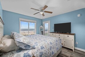A bedroom with a bed, a ceiling fan, a television, and a view of the beach.