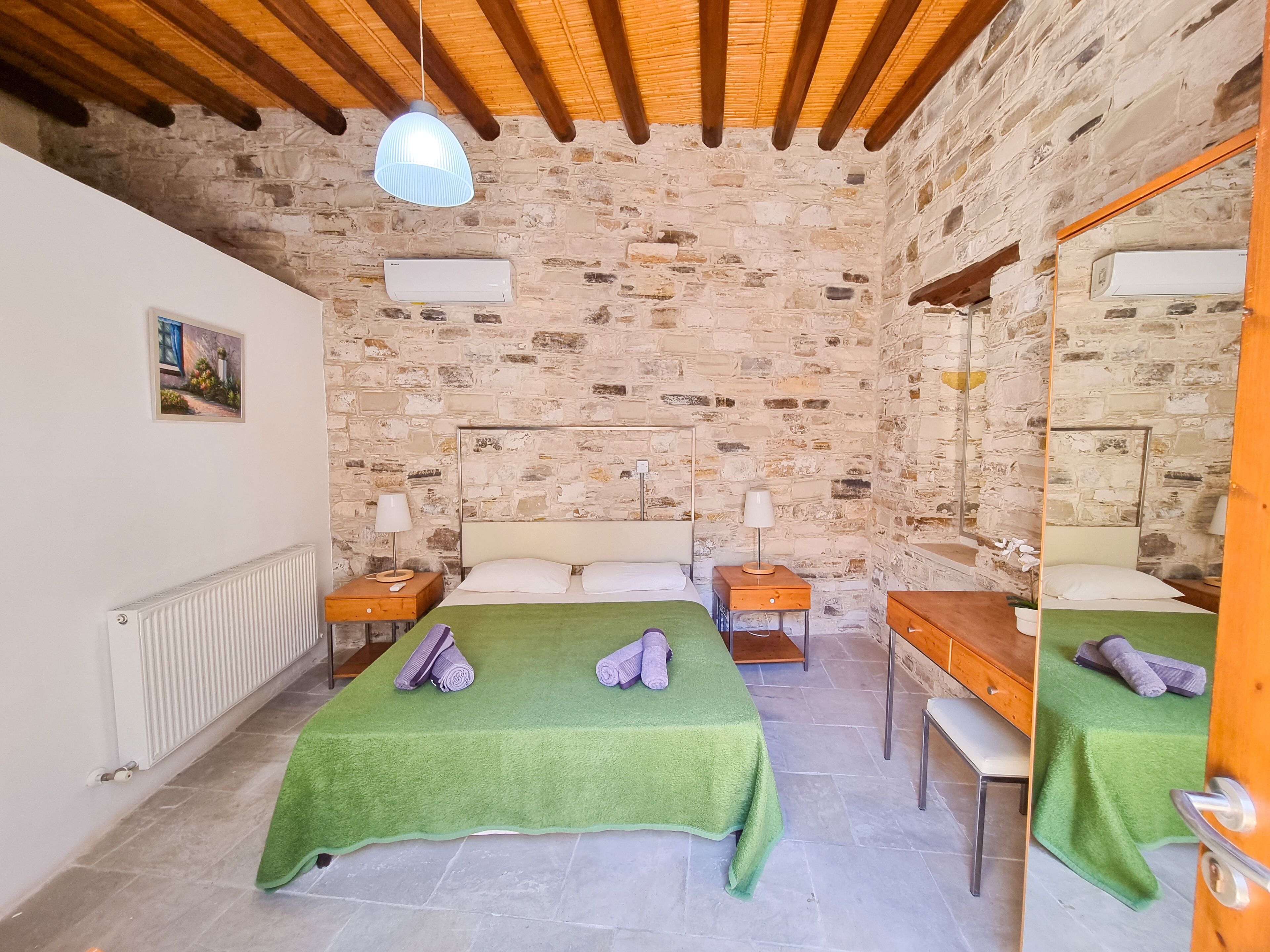 A room with stone walls, wooden beams, a bed with green bedding, a wooden nightstand, a mirror, and a radiator.