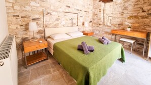 Une chambre avec un mur en pierre, un lit recouvert d’une couverture verte, une table de chevet en bois, une lampe sur la table de chevet et un miroir au-dessus du bureau.