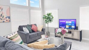 Un salon moderne avec un canapé gris, des coussins colorés, une table basse ornée d’un vase de fleurs et un téléviseur à écran plat posé sur un meuble en bois.
