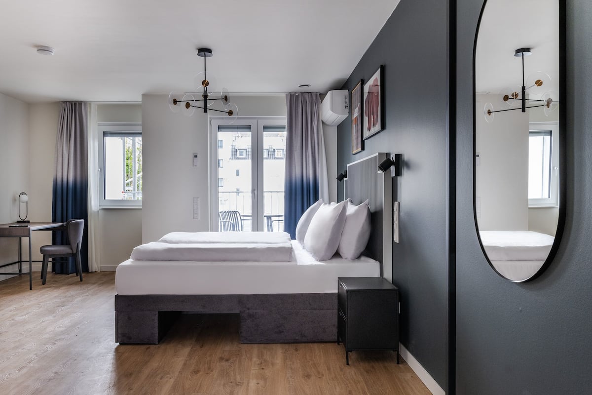 Quarto moderno com uma cama grande, uma escrivaninha e uma cadeira. Há uma janela com cortinas, uma luminária no teto e um espelho fixado na parede.