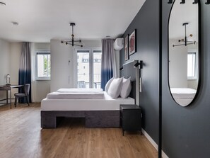 A modern bedroom with a large bed, a desk, and a chair. There is a window with curtains, a ceiling light, and a wall-mounted mirror. - Numa Düsseldorf Leo (Düsseldorf)