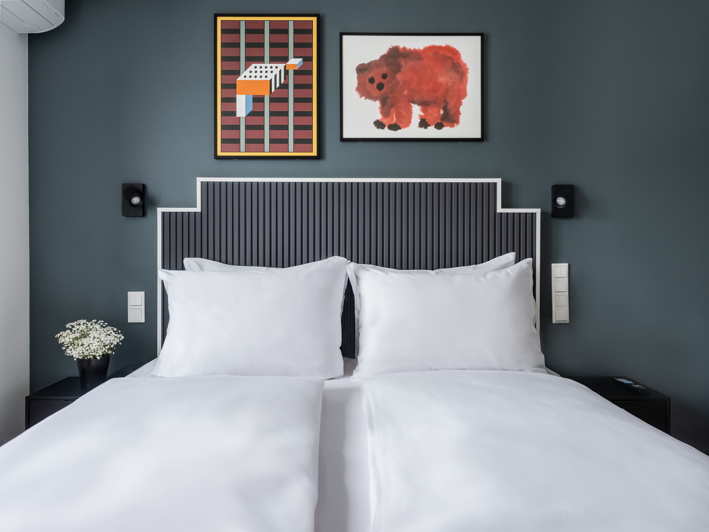 Een net opgemaakt bed met witte lakens, een gestreept hoofdbord en twee lichtschakelaars aan de muur.