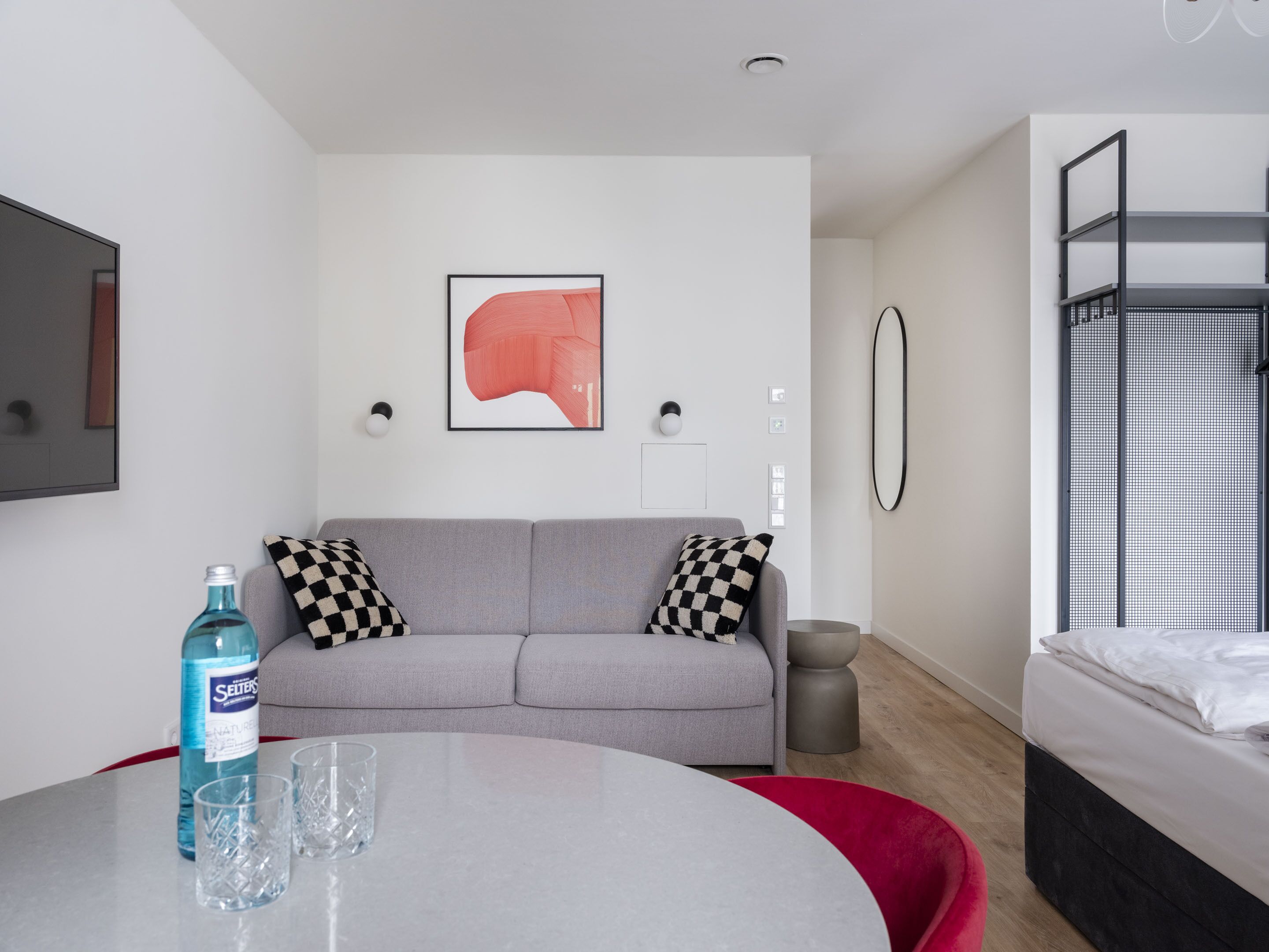 A modern, minimalist room with a grey sofa, a round table with a bottle and glasses, a bunk bed, and a framed abstract artwork on the wall.