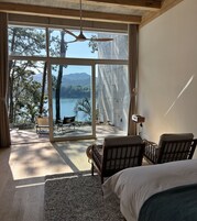 Un dormitorio con un ventanal grande, ventilador de techo y vista a un lago y árboles.