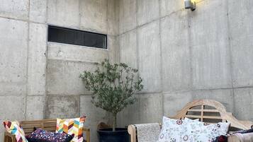 Ein gemütliches Zimmer mit einer Betonwand, einer hölzernen Bank, einem hölzernen Bett, einer Topfpflanze und zwei hängenden Korbstühlen.