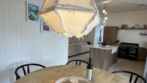 Ruokailutilassa on pyöreä puinen pöytä, kudottu alusta ja pullo viiniä. Taustalla näkyy keittiö, jossa on kalanruotokuvioinen välitila ja liesi.