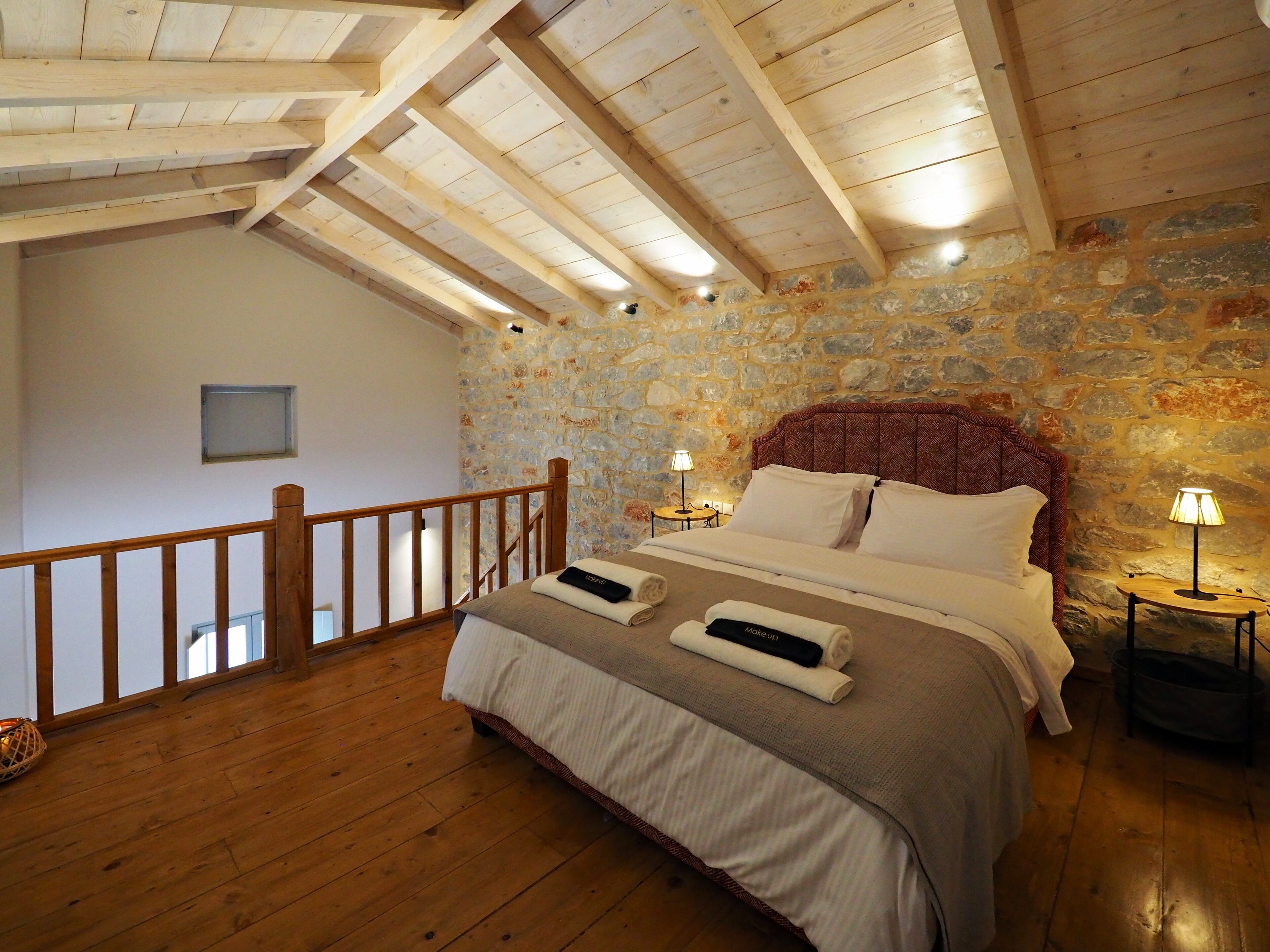 Un dormitorio con pared de piedra, vigas de madera en el techo, una cama con sábanas blancas y dos lámparas de noche.