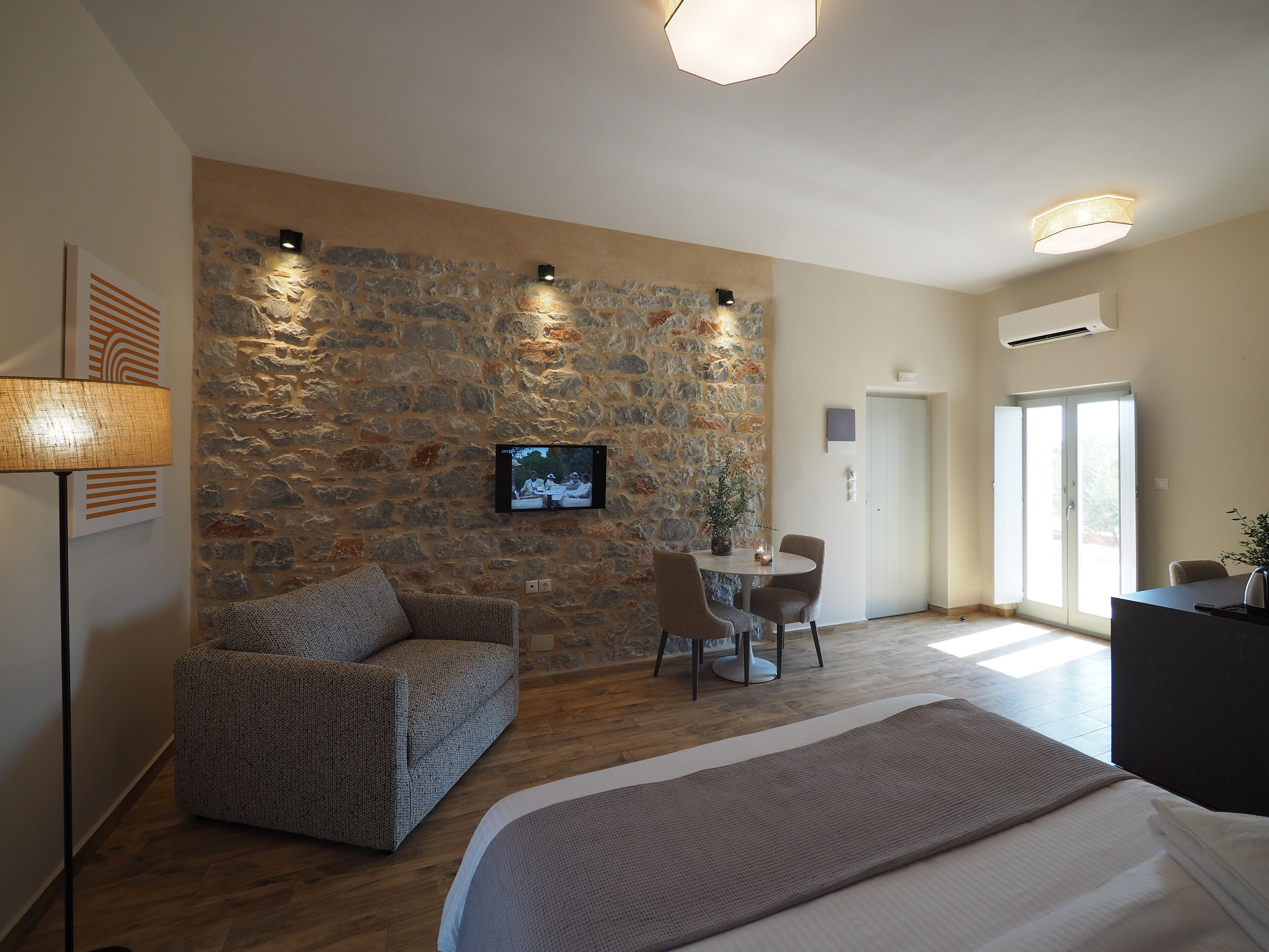 Une chambre moderne avec un mur en pierre, un lit, un coin salon avec une table et un téléviseur fixé au mur.