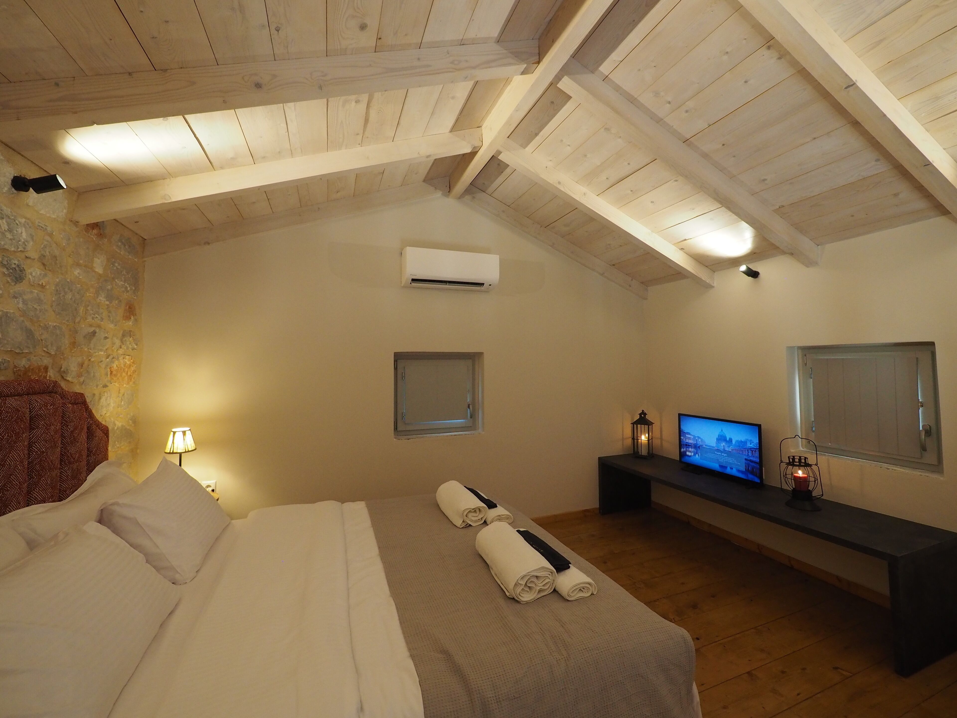 Ein Schlafzimmer mit Holzbalkendecke, einem Bett mit weißer Bettwäsche, einem kleinen Tisch mit Fernseher und einer Steinwand.