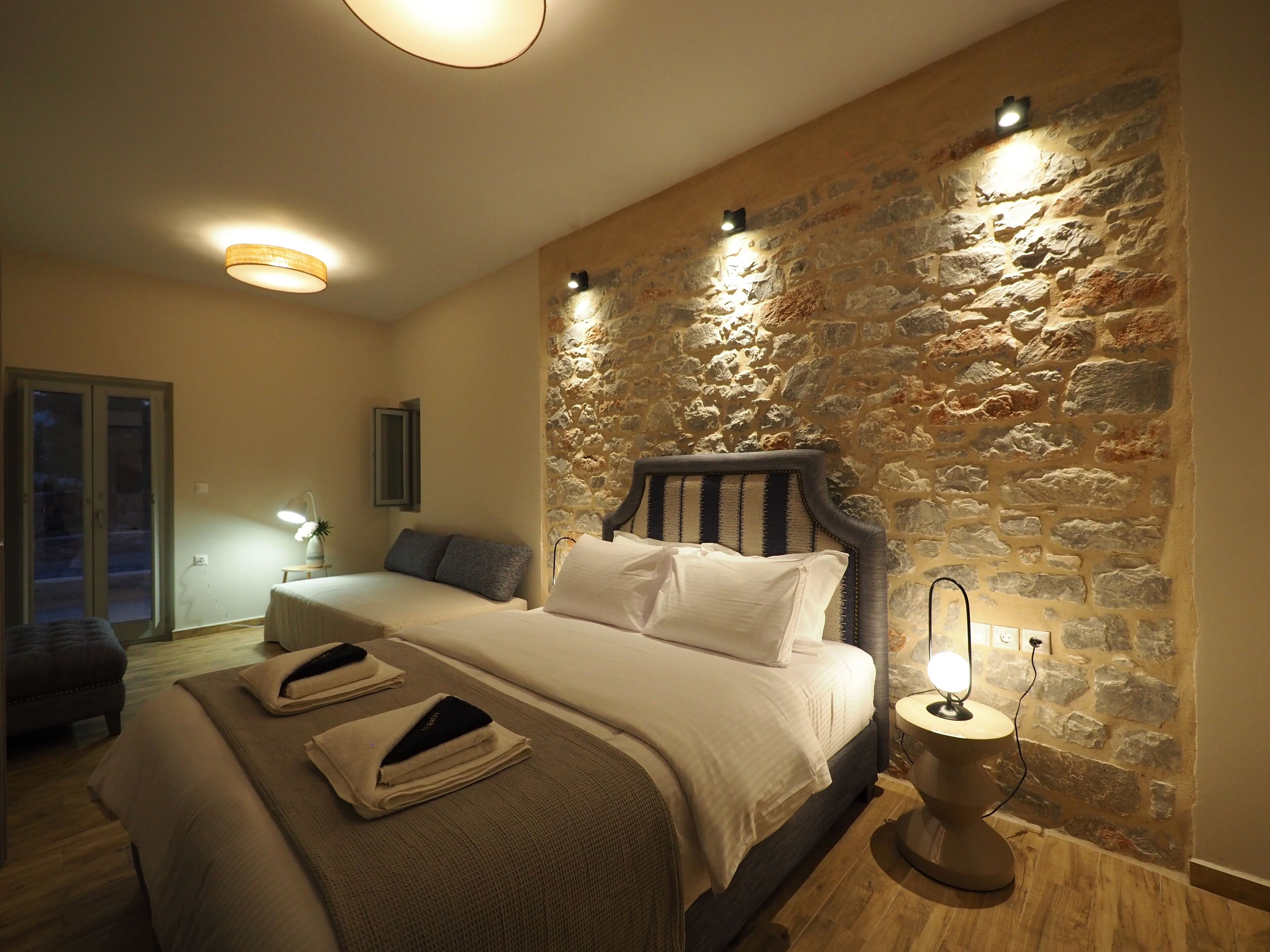 Ein Schlafzimmer mit Steinswand, einem Bett mit Kissen, einem Nachttisch mit Lampe und Blick durch eine Tür.
