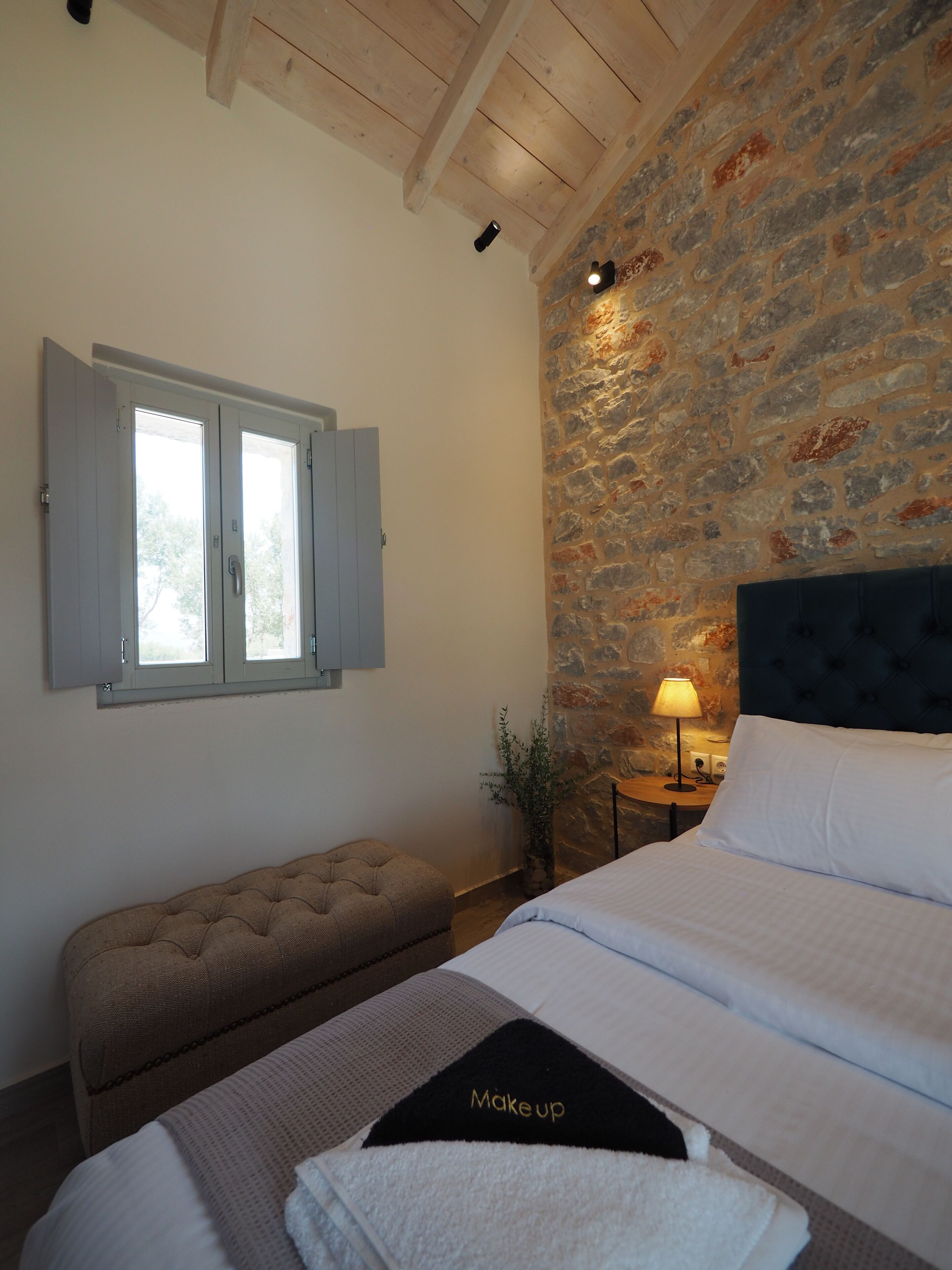A bedroom with a stone wall, a bed with a 'Makeup' pillow, a bench, and a nightstand with a lamp.
