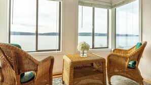 A room with two wicker chairs, a small table with a vase of flowers, and large windows overlooking a body of water.