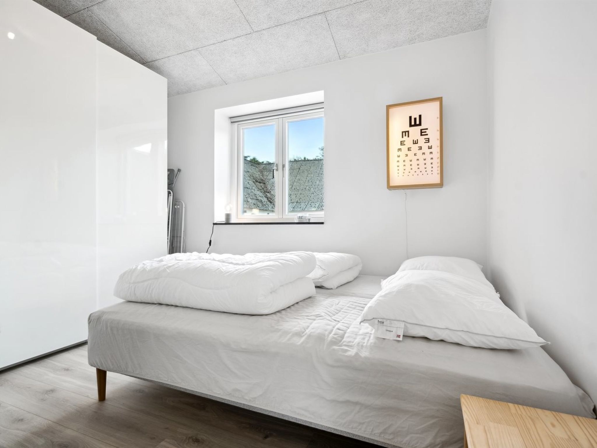 Une chambre à coucher minimaliste, avec un lit, une fenêtre et un tableau d’acuité visuelle fixé au mur.