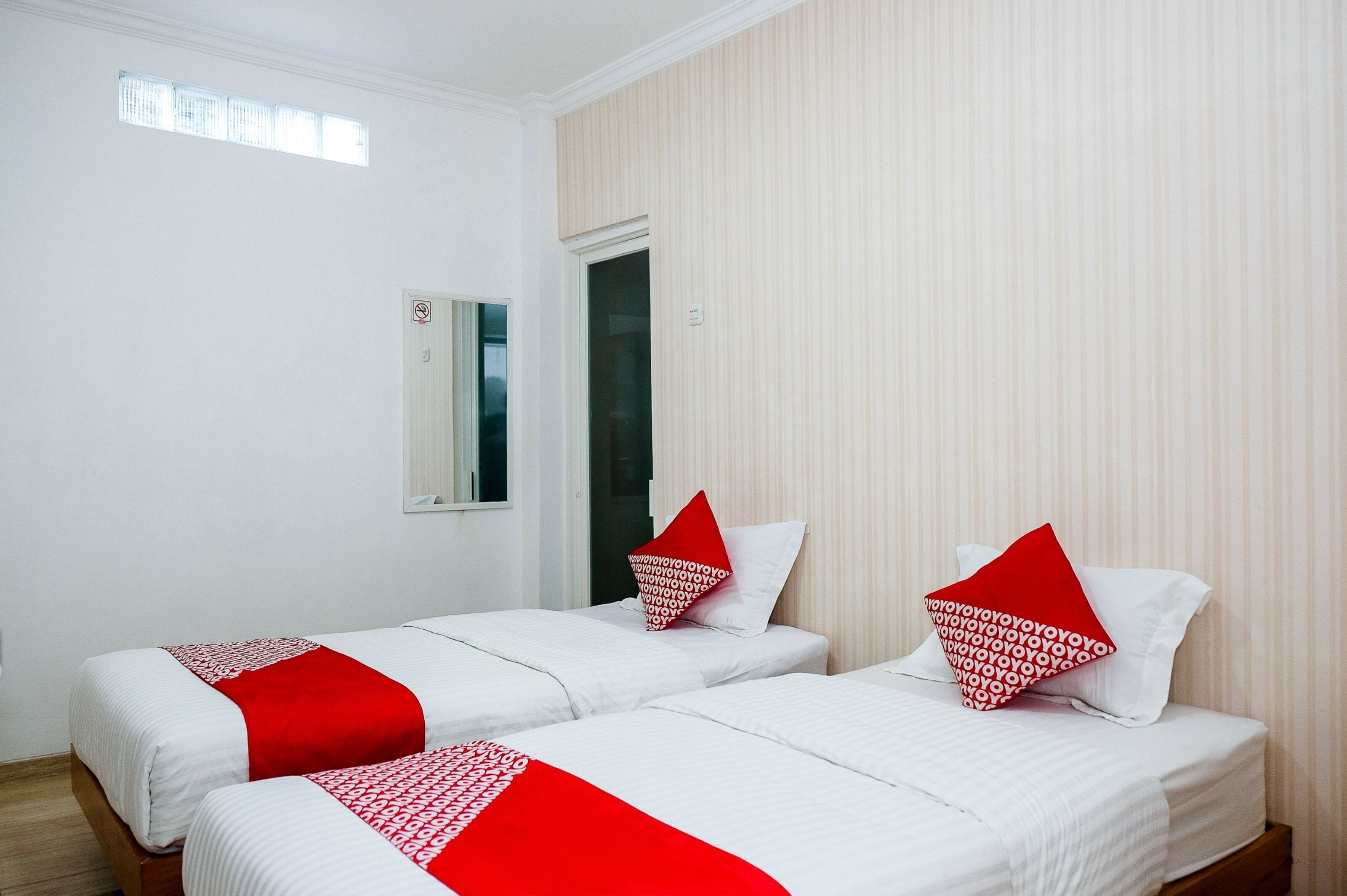 A hotel room with two single beds, each with white bedding and red accents, a window, and a door.