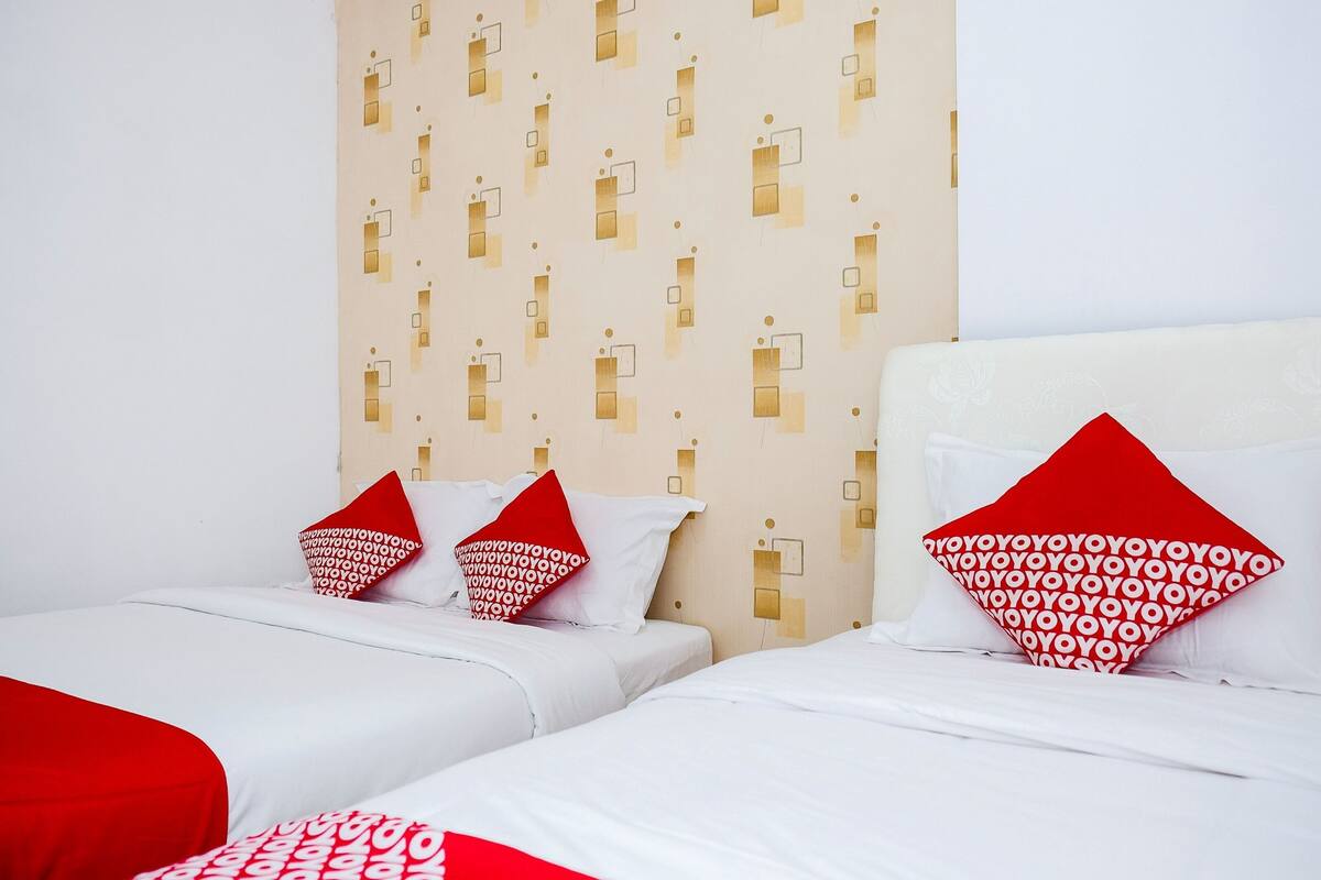A hotel room with two beds, each with white bedding and red pillows. The room has a patterned wall and a plain wall with a light switch design.