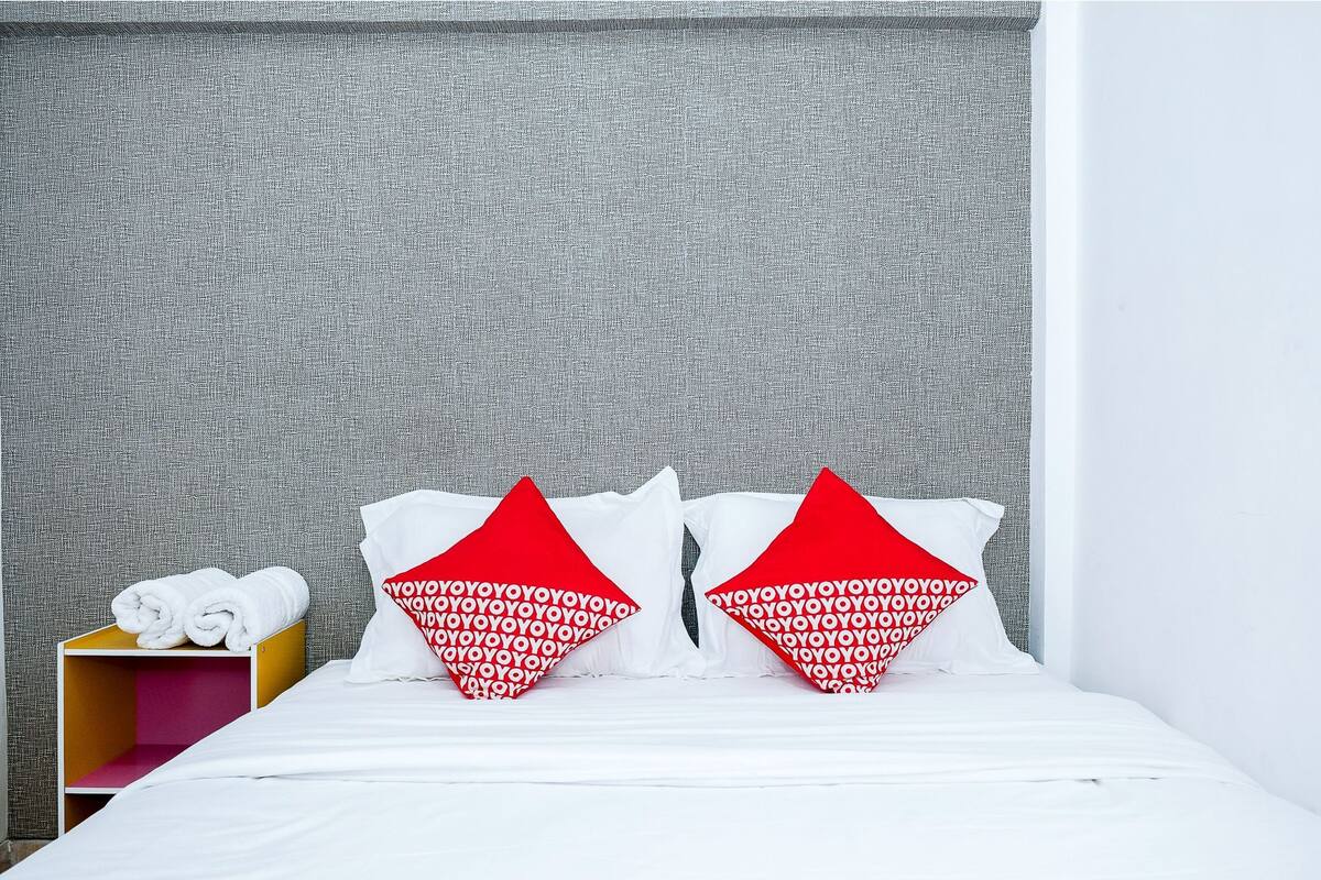 A hotel room with a neatly made bed, two pillows with a red and white pattern, a small wooden nightstand with rolled towels, and a grey textured wall.