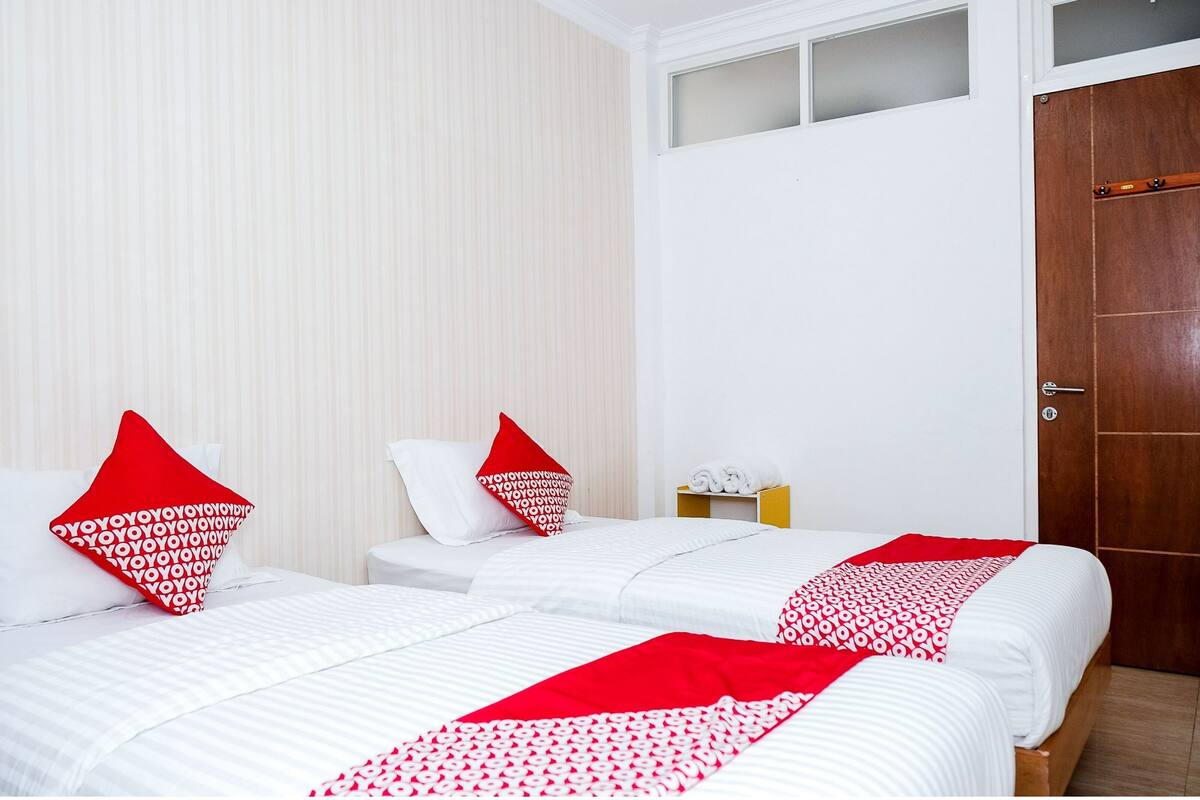 A hotel room with two beds, each with white bedding and red pillows. There is a wooden door and a small table with towels.