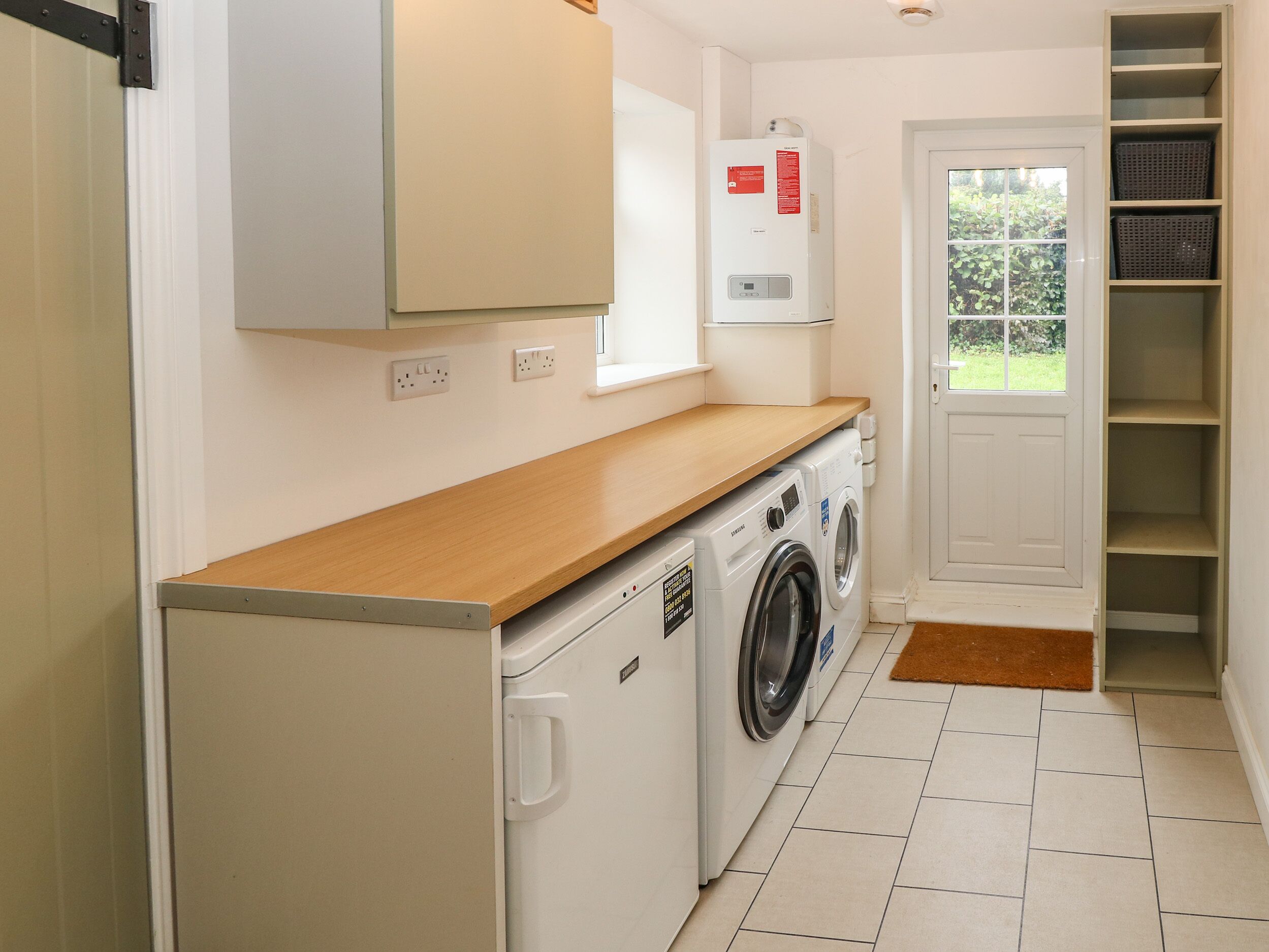 A compact laundry room with a washing machine, dryer, and a water heater. There is a wooden countertop and a door leading to a garden.