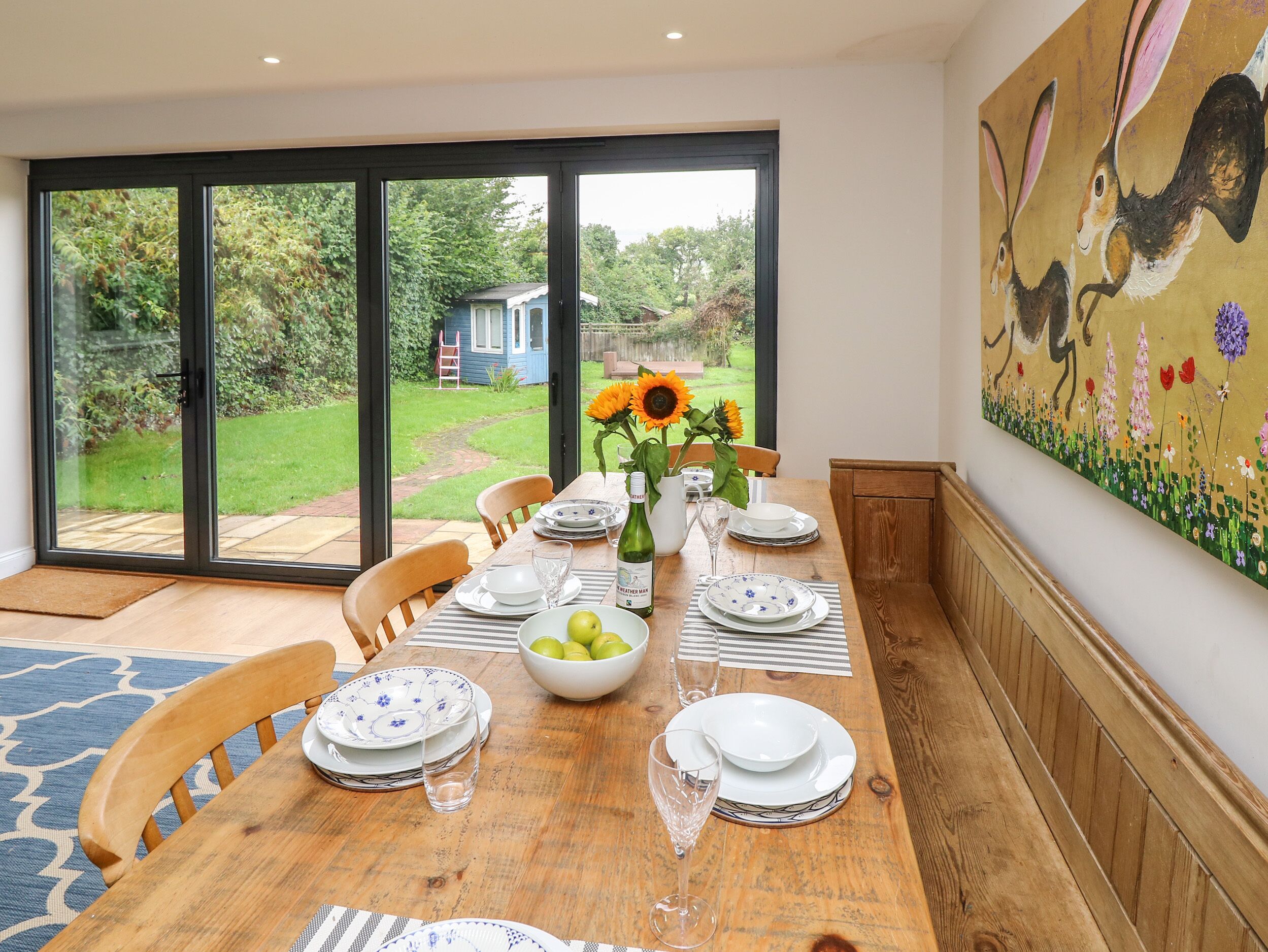 A dining area with a table set for four, large windows, and a painting on the wall.