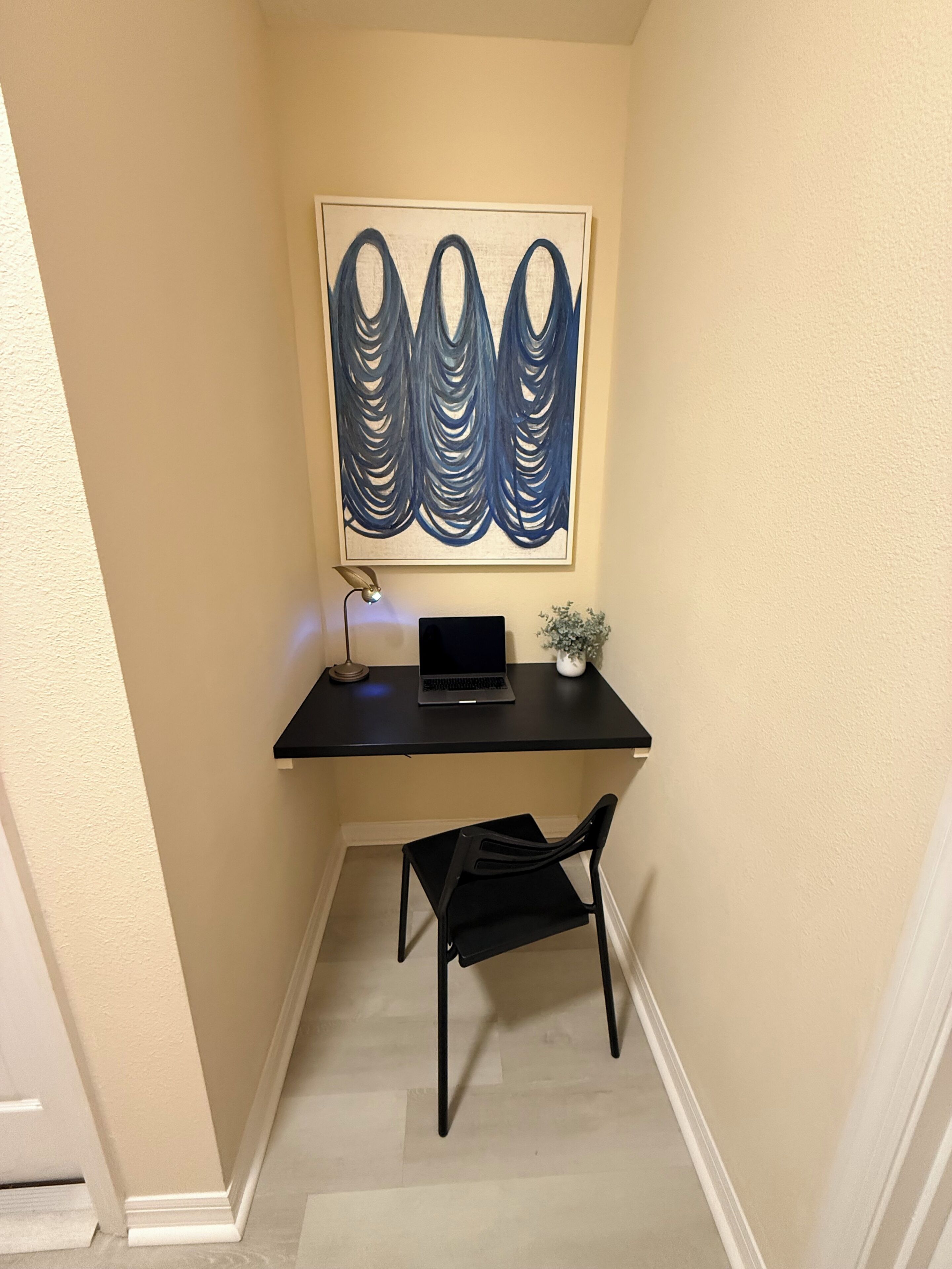 A small workspace with a desk, laptop, and chair, featuring a wall-mounted abstract painting.