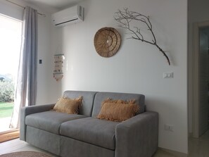 A living room with a grey sofa, two throw pillows, a wall-mounted air conditioner, a wall art piece, and a macrame wall hanging. - Apartment With sea View (Pachino)