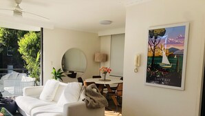 Una sala moderna con un sofá blanco, un espejo redondo, una zona de comedor con mesa y sillas, y un ventanal con vistas a zonas verdes.
