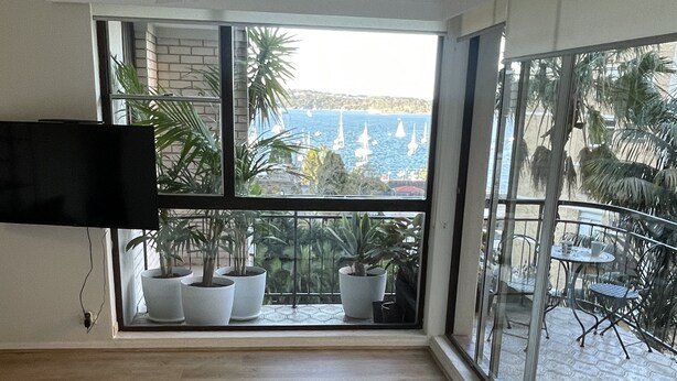 Una sala de estar moderna con un sofá grande, un televisor de pantalla plana montado en la pared y un balcón con plantas en macetas y vistas al mar.