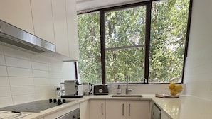 Una cocina moderna con gabinetes blancos, una campana extractora de acero inoxidable, horno integrado, lavavajillas y una ventana con vistas a árboles.