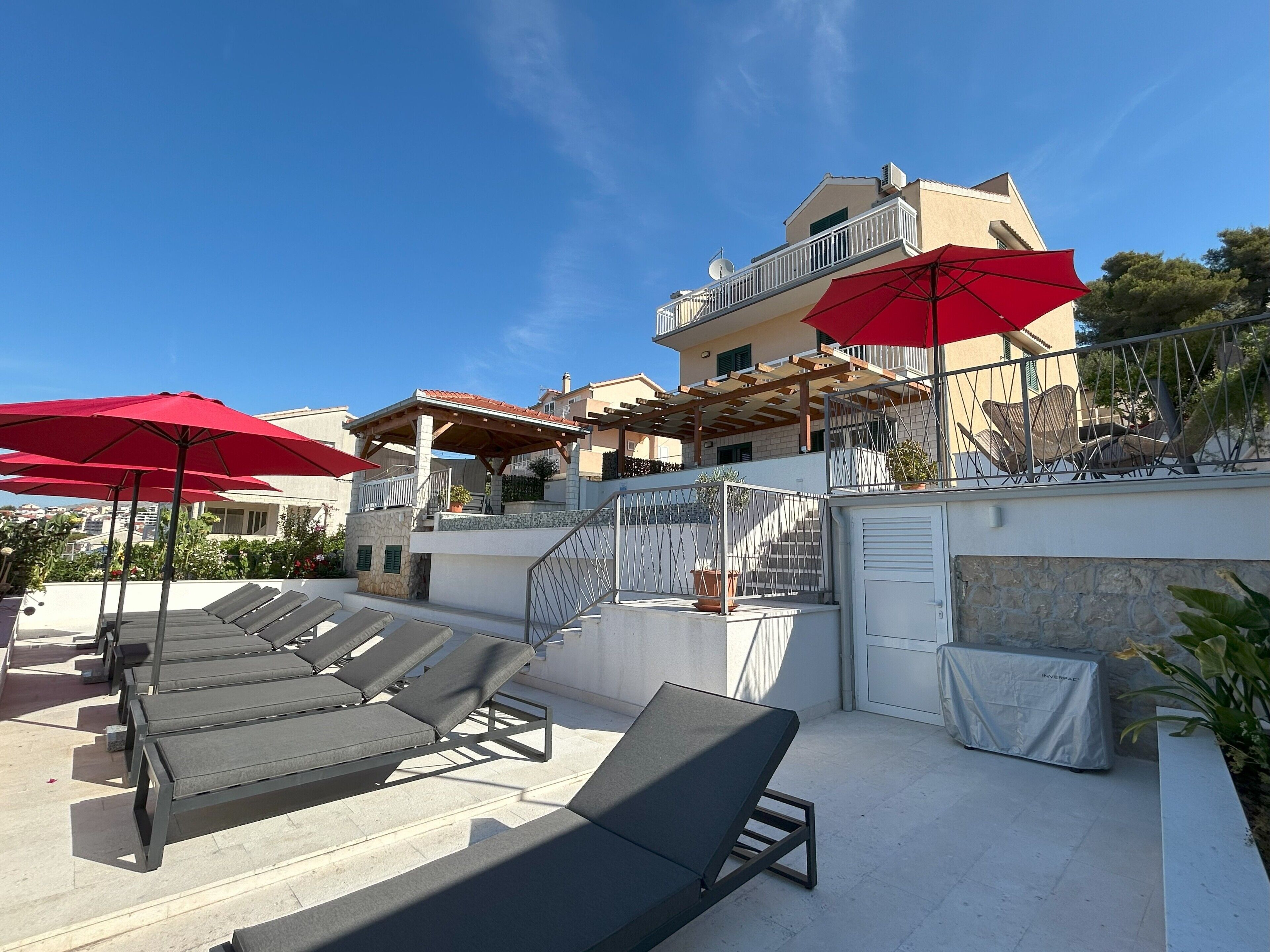 A rooftop terrace with sun loungers and red umbrellas, a stone wall, and a building with a balcony in the background.