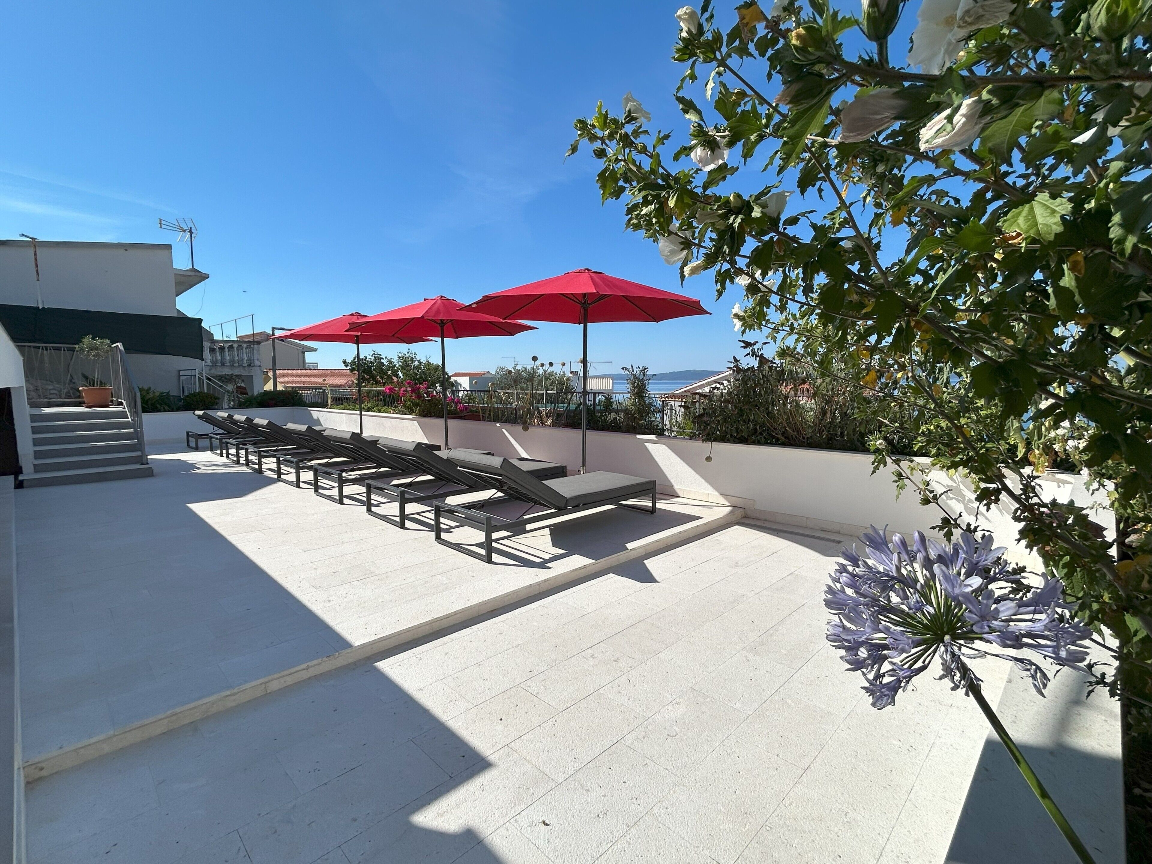 A rooftop terrace with sun loungers and red umbrellas, a staircase, and a view of the sea.