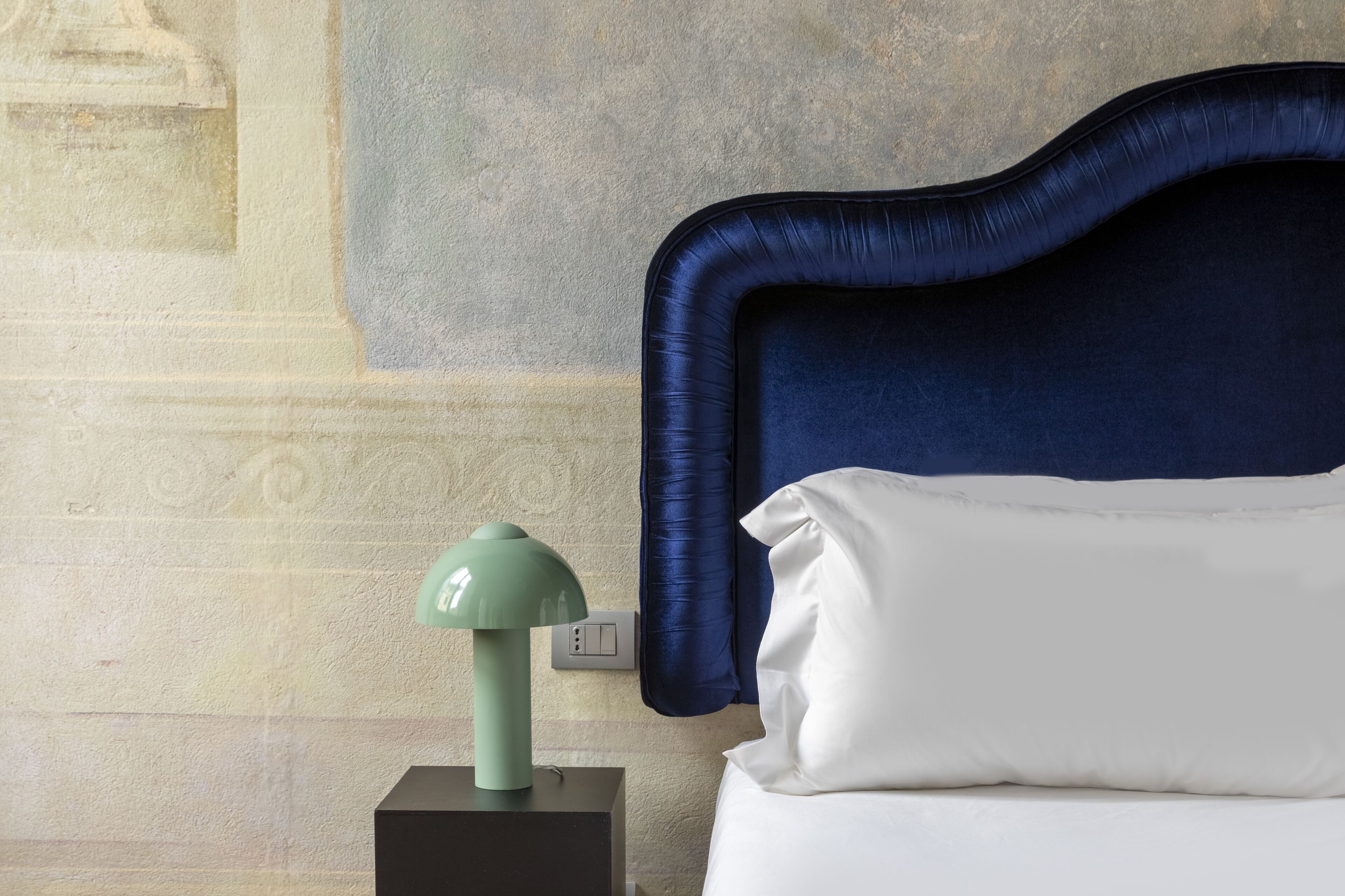 A neatly made bed with a white pillow and a dark blue headboard. A green lamp is placed on a black nightstand beside the bed.