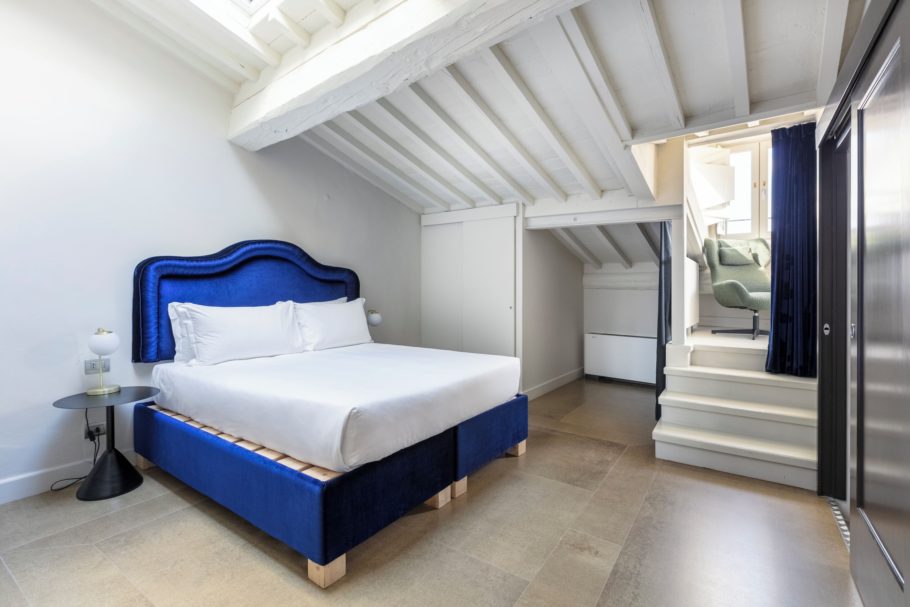 Quarto com uma cama, mesa de cabeceira, abajur e escada que leva a outro cômodo.