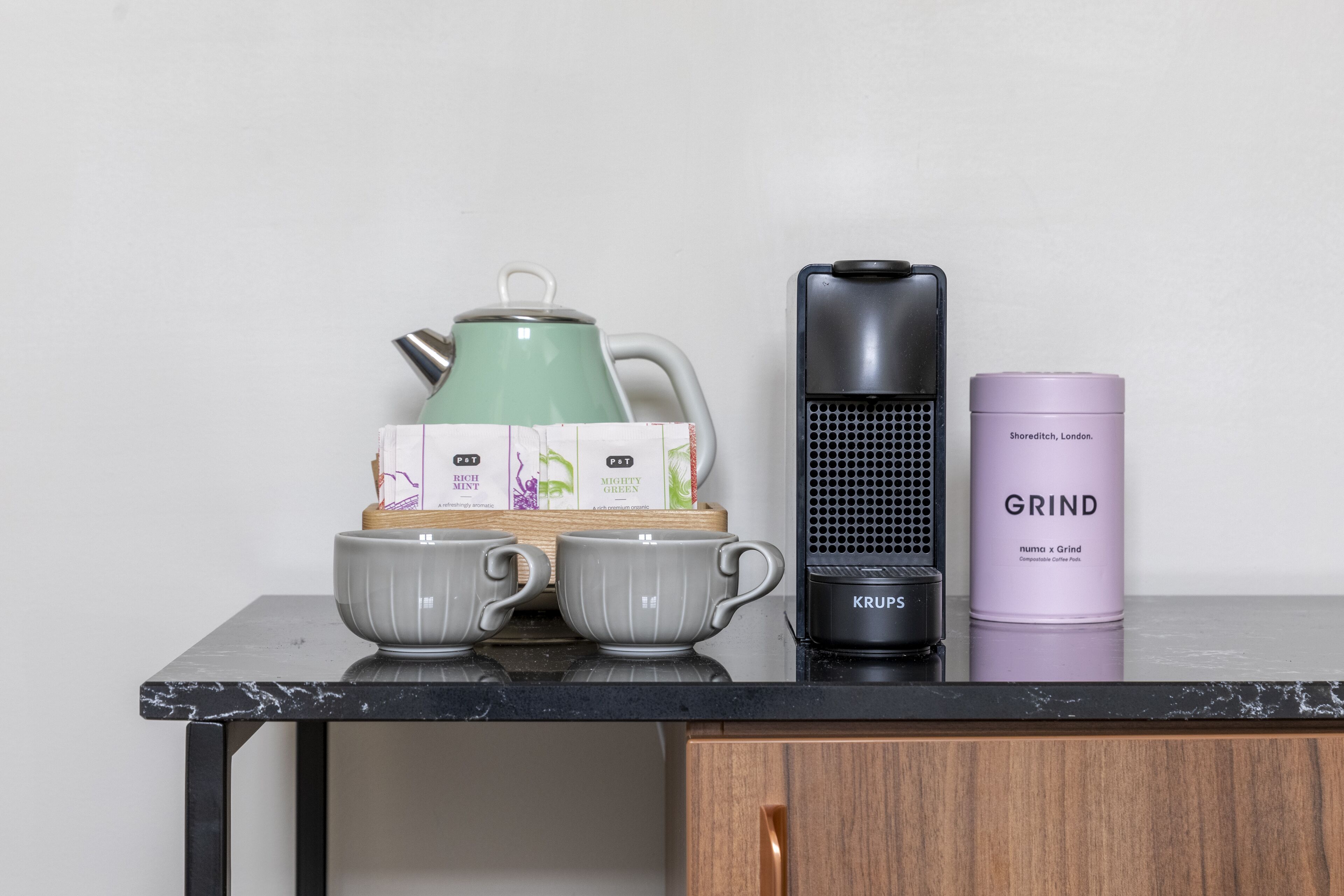 Una encimera de cocina con un hervidor verde, dos tazas grises, una cafetera Krups y un recipiente púrpura con la etiqueta 'Shoreditch, London'.