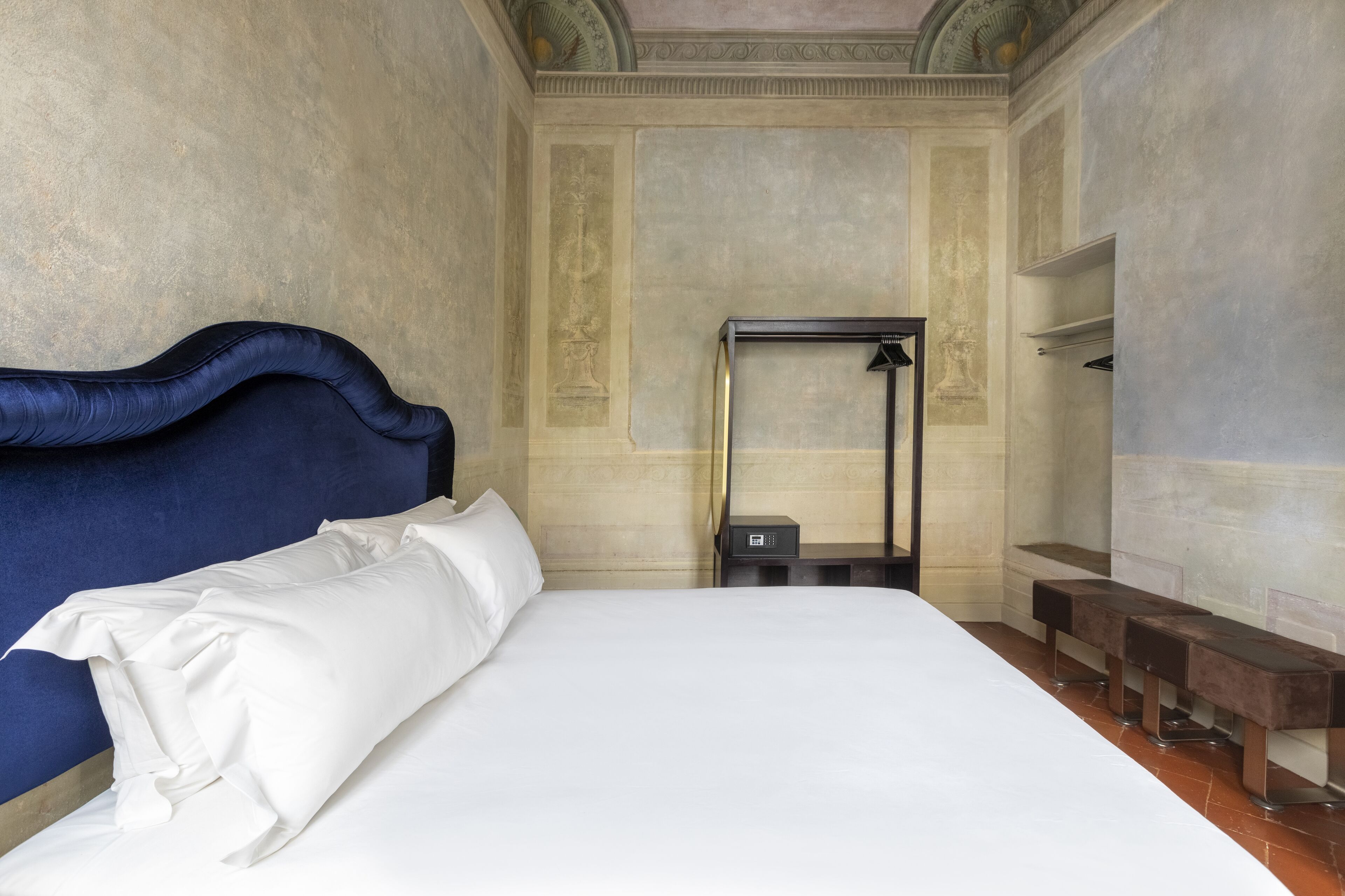 A hotel room with a large bed, a headboard, a bedside table, a mirror, and a bench along the wall.