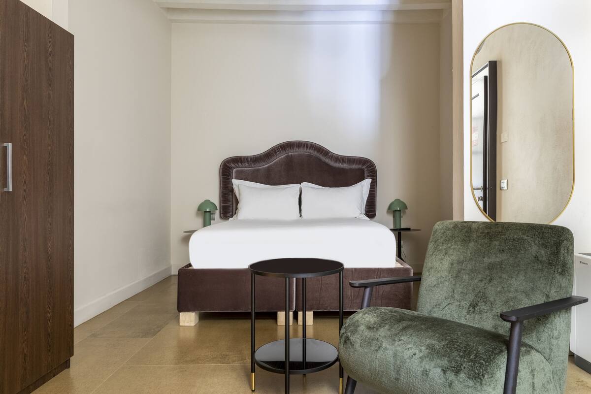 A bedroom with a bed, a round side table, a green chair, a wooden wardrobe, and a mirror.