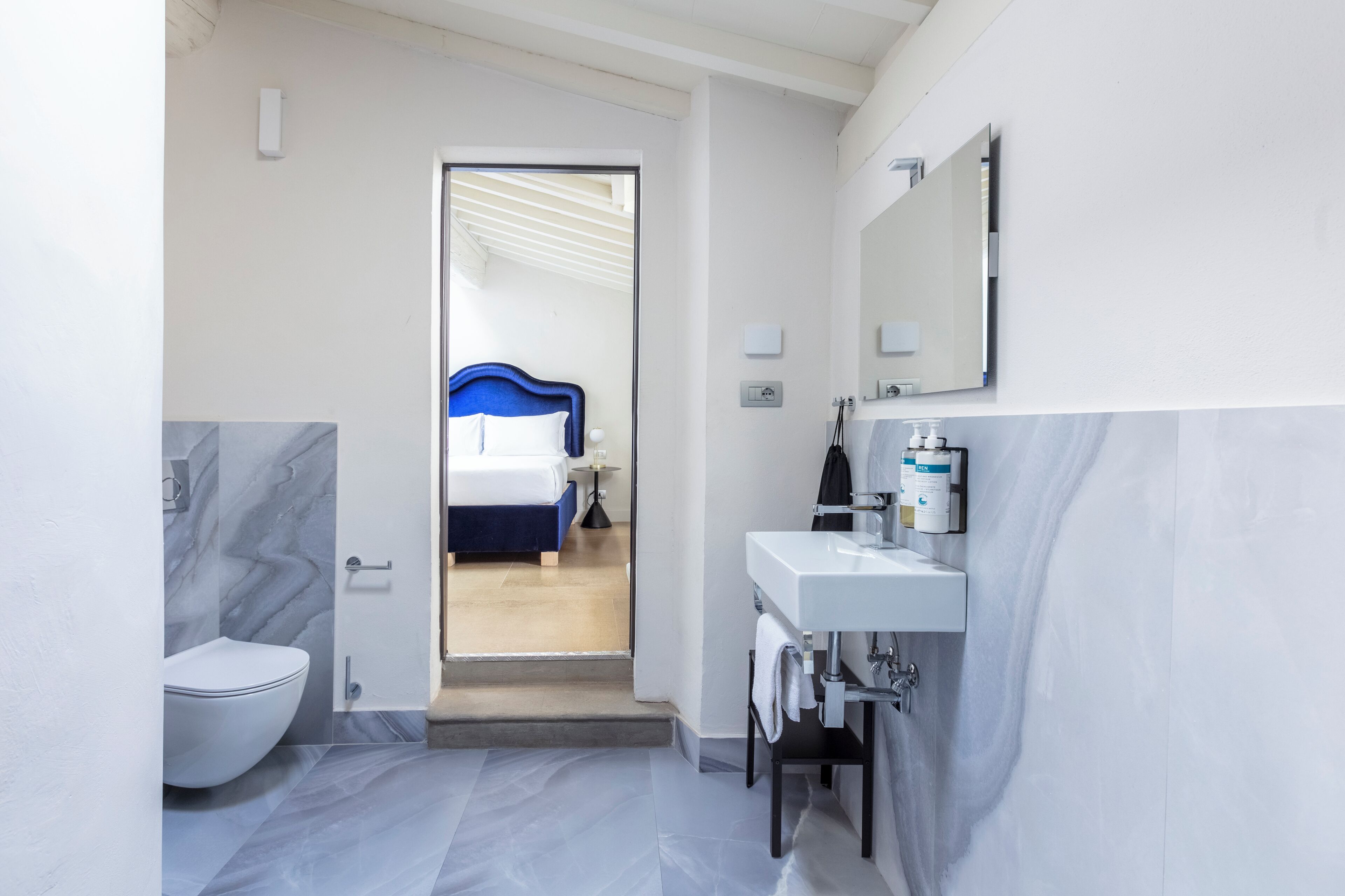 A modern bathroom with a white toilet, marble flooring, a blue headboard bed, and a white sink with a mirror.