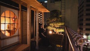 Une structure traditionnelle japonaise en bois, dotée d’une fenêtre circulaire, d’un petit banc en bois et d’un arbre bonsai.