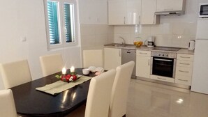 A modern kitchen with white cabinets, a dining table with chairs, and a window with blinds. - Heated infinity pool, family apartment with panoramic sea view 2 (Okrug Gornji)