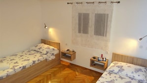 A small room with two wooden beds, a window with curtains, a nightstand, and a wall-mounted lamp. - Heated infinity Pool, family apartment with panoramic sea view 3 (Okrug Gornji)