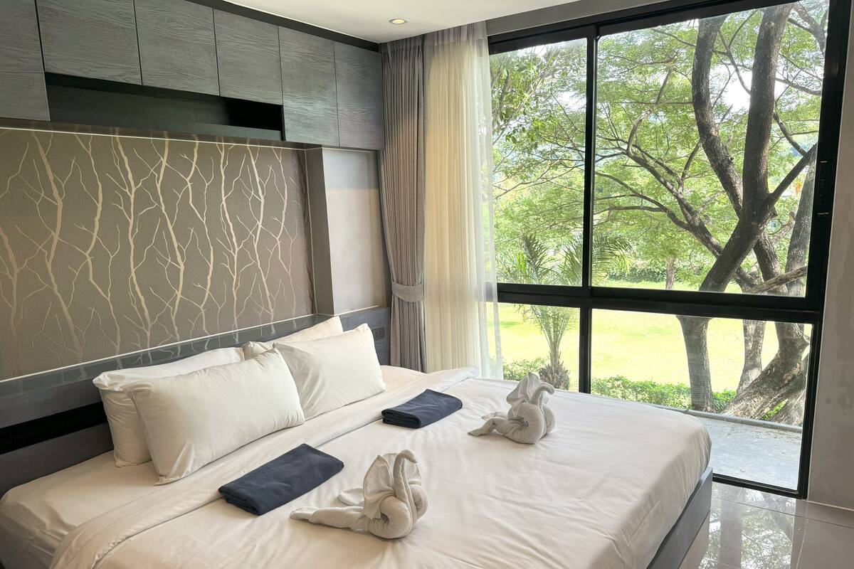 A neatly made bed with white linens and two towels, a headboard with a tree branch pattern, and a large window with a view of trees and grass.
