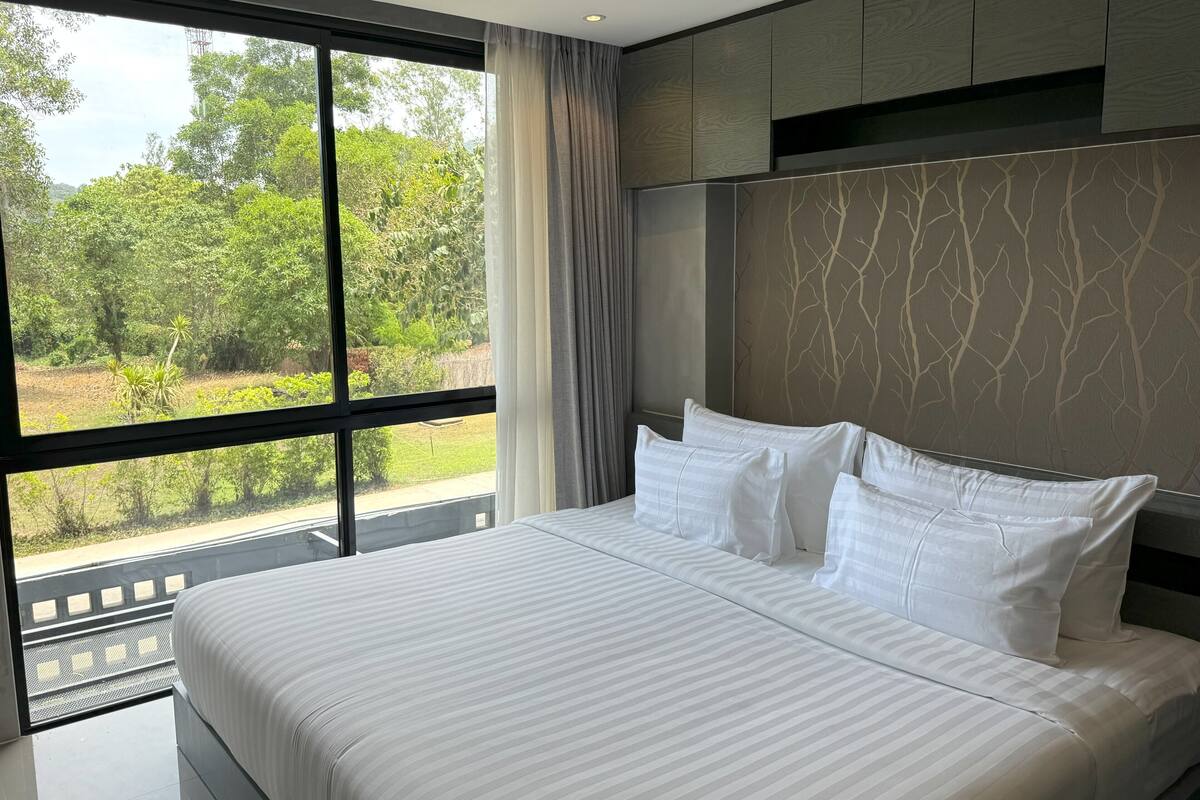 A modern hotel room with a large window, a bed with white linens, and a view of greenery outside.