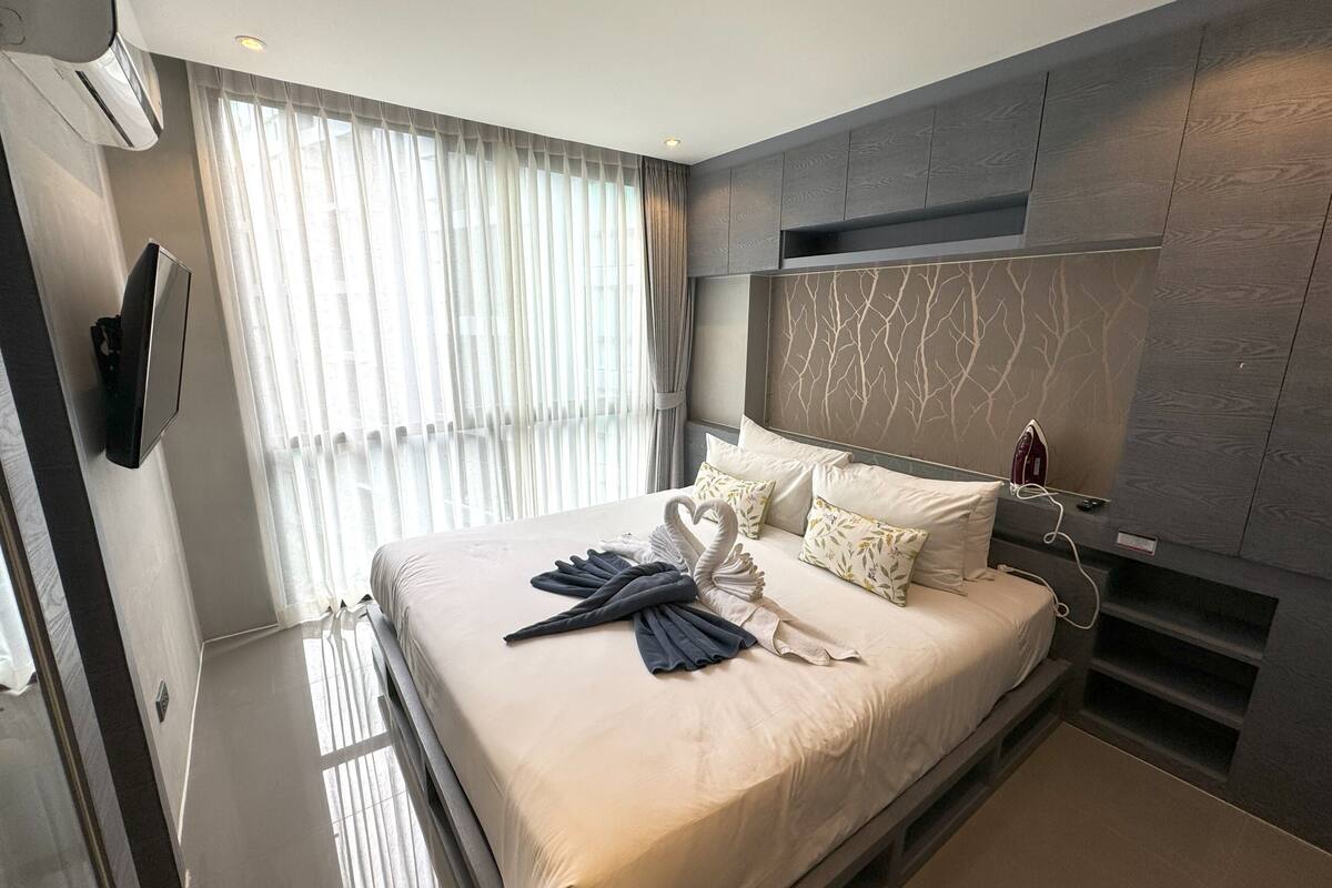 A modern hotel room with a large bed, a flat-screen TV, a built-in wardrobe, and a large window with sheer curtains.