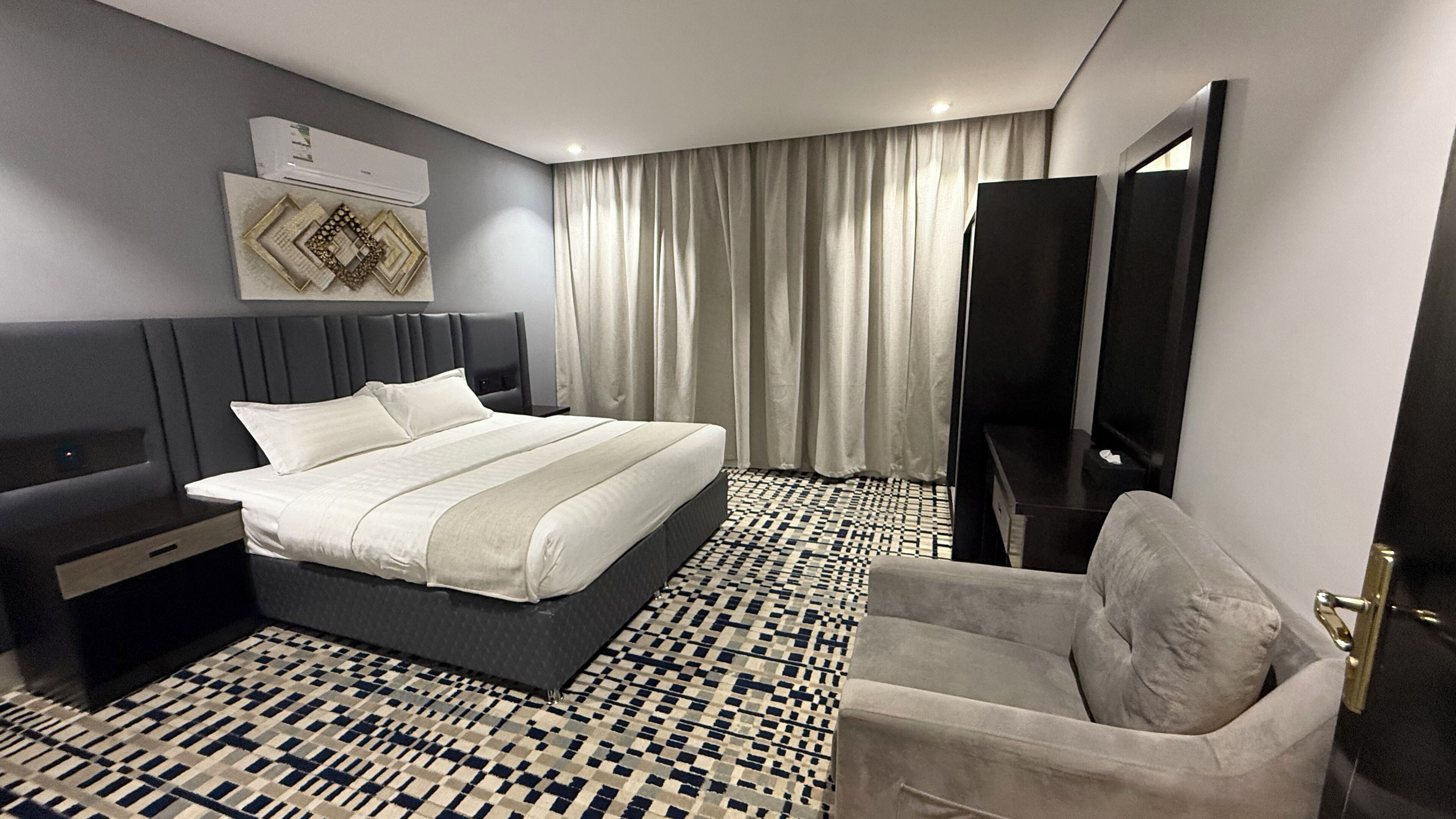 Habitación de hotel moderna con una cama grande, una mesita de noche, una silla, un televisor y un suelo estampado.