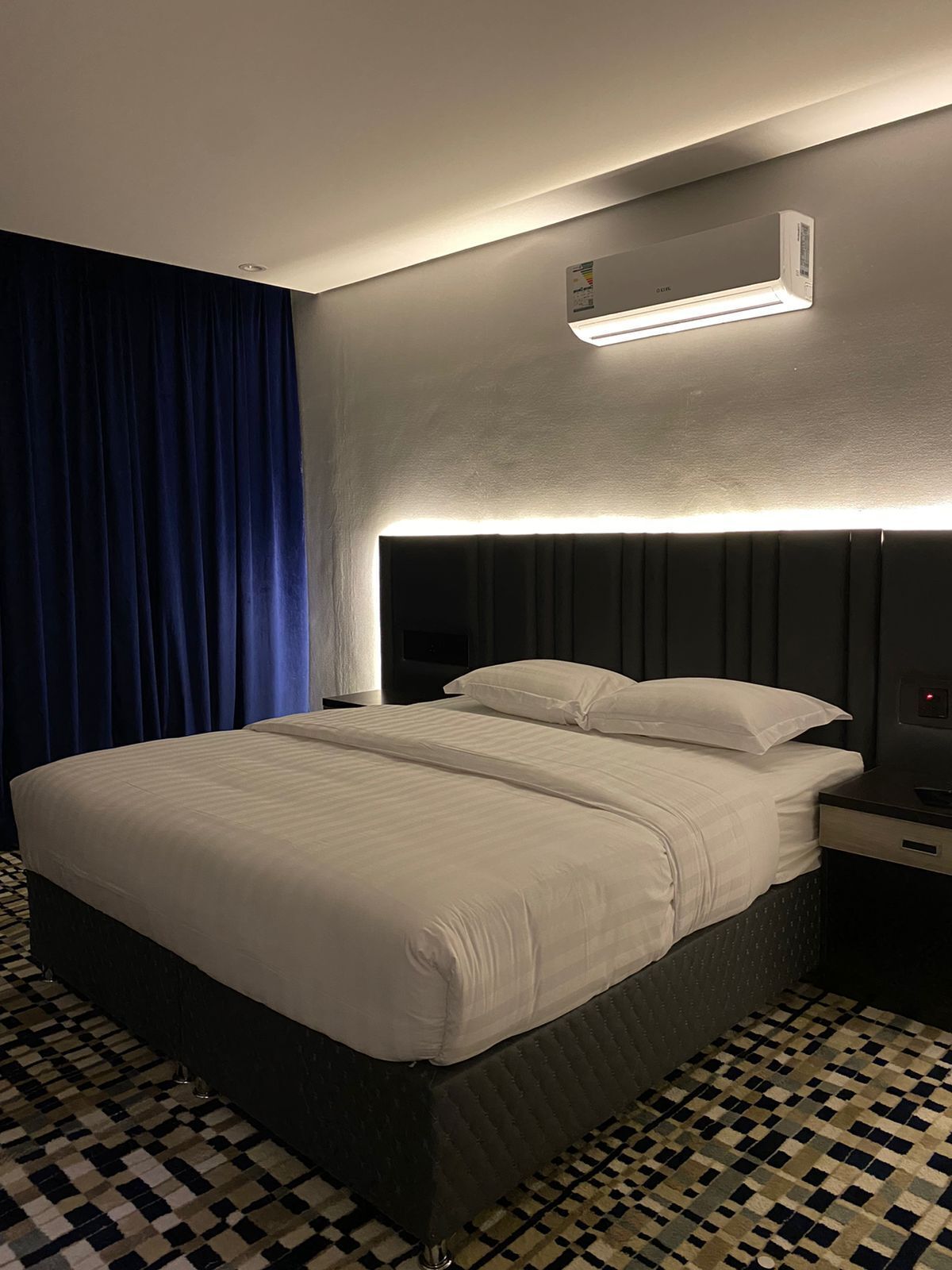 Une chambre d’hôtel avec un grand lit, une tête de lit dotée d’un éclairage intégré, une table de chevet avec une lampe et un motif de carreaux au sol.