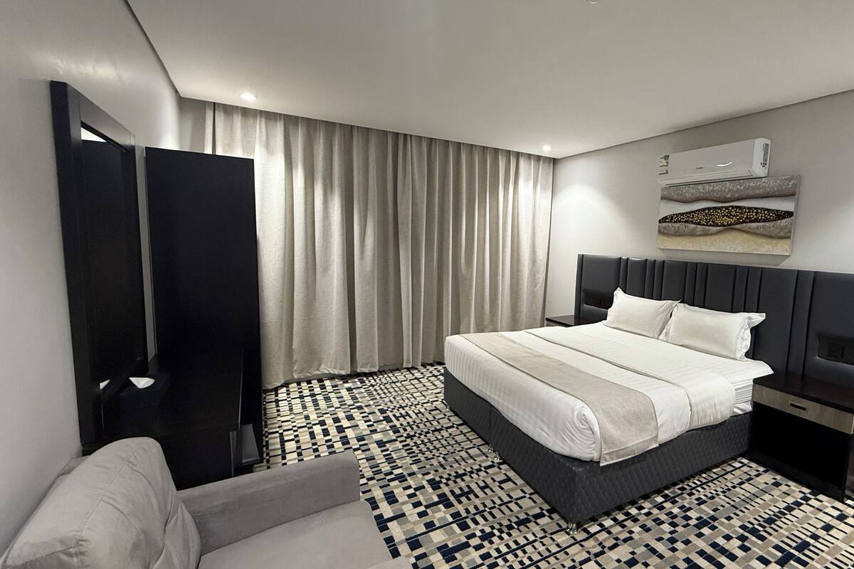 Une chambre d’hôtel moderne avec un grand lit, une table de chevet, une chaise, un tapis à motifs et une grande fenêtre avec des rideaux.